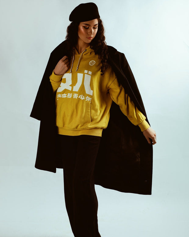 Person wearing a yellow hoodie with white text and a black coat on a plain background